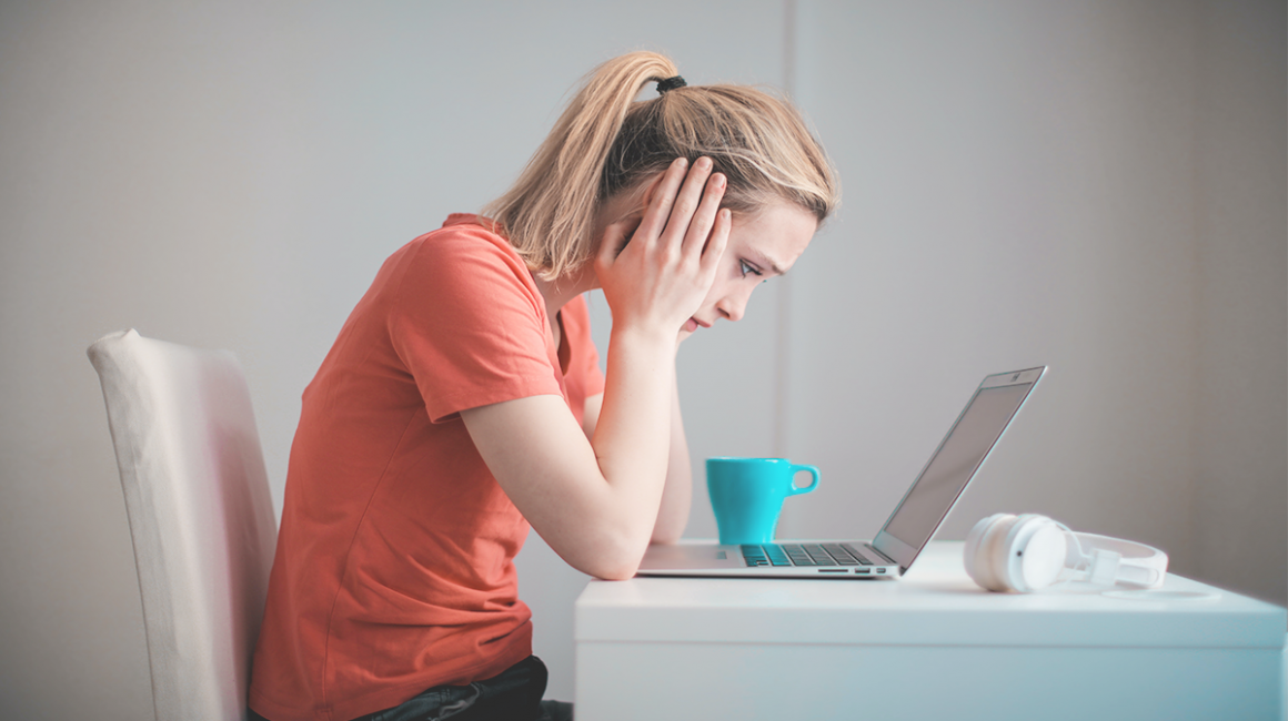Dziewczyna ukazana bokiem, która siedzi przy biurku nad laptopem. Ma zatroskaną twarz. Łokcie opiera na blacie. Głowę ma opartą na dłoniach.