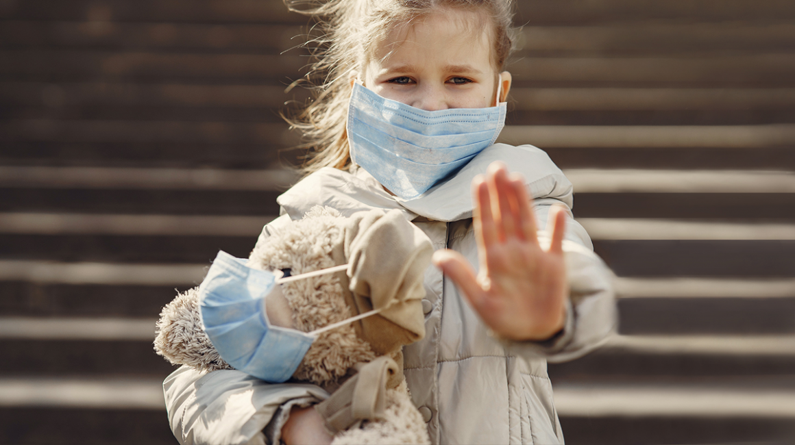 Dziewczynka z misiem pod ręką, mają założone maseczki. Lewa ręka dziewczynki wyciągnięta w przód, otwartą dłonią.