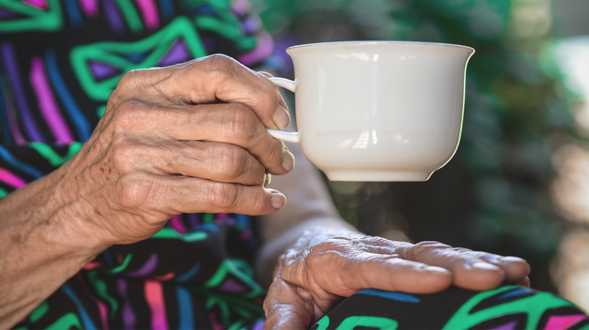 Wykadrowana starsza kobieta, która siedzi i w prawej ręce trzyma filiżankę. Widać ręce, filiżankę i fragment tułowia.