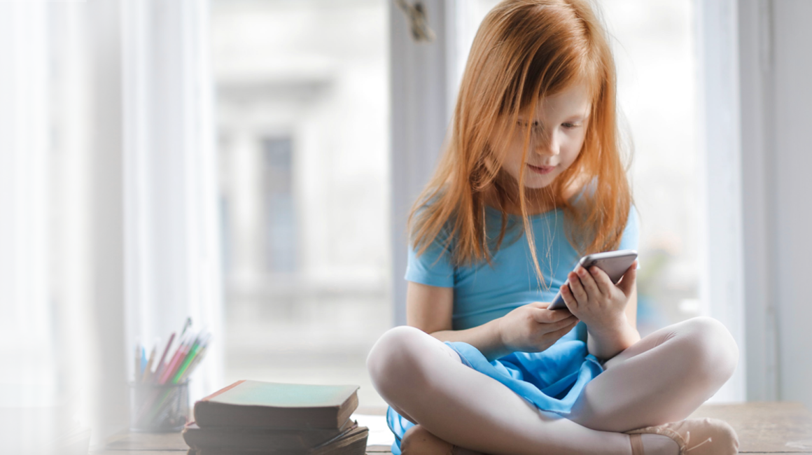 Dziewczynka siedząca ze skrzyżowanymi nogami na tle okna, przeglądająca telefon komórkowy.