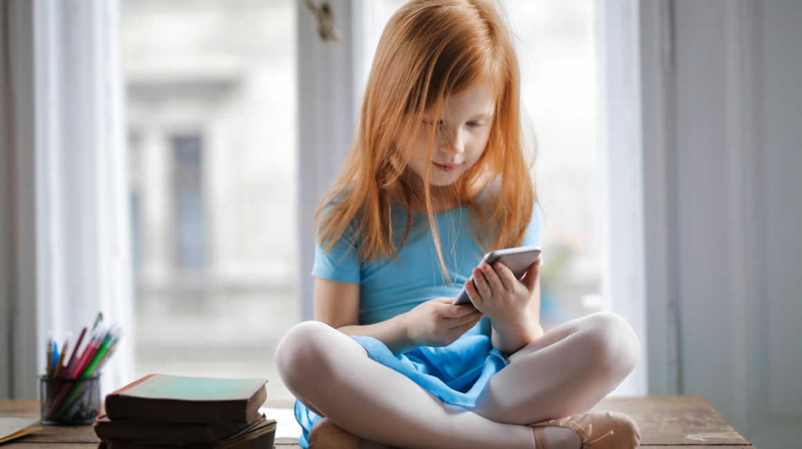 Dziewczynka siedząca ze skrzyżowanymi nogami na tle okna, przeglądająca telefon komórkowy.