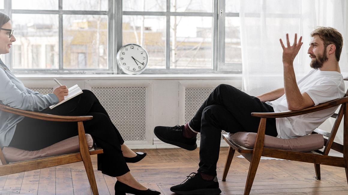 Mężczyna i kobieta siedzący naprzeciwko siebie. Kobieta ma notes i długopis. Mężczyzna opowiada z gestykulacją 