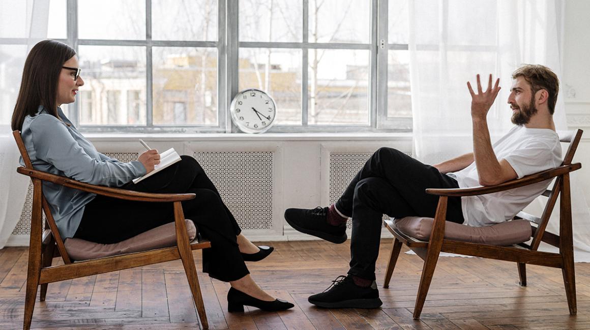 Mężczyna i kobieta siedzący naprzeciwko siebie. Kobieta ma notes i długopis. Mężczyzna opowiada z gestykulacją 