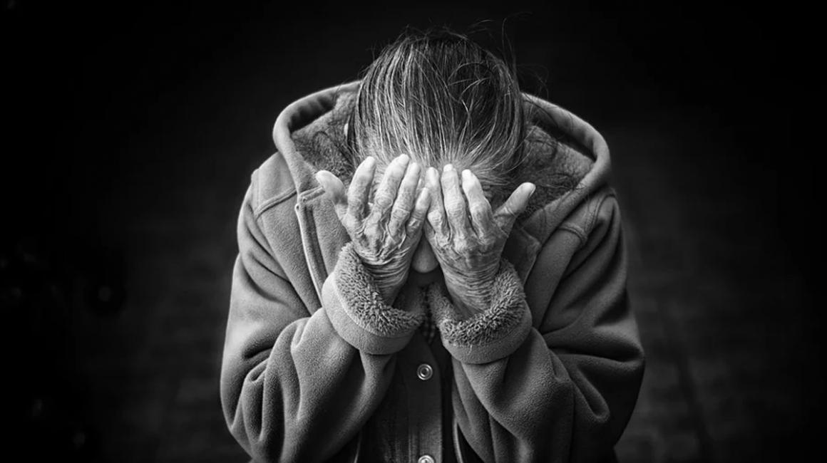 Starsza kobieta pochylona, z rękoma zasłaniącymi twarz.