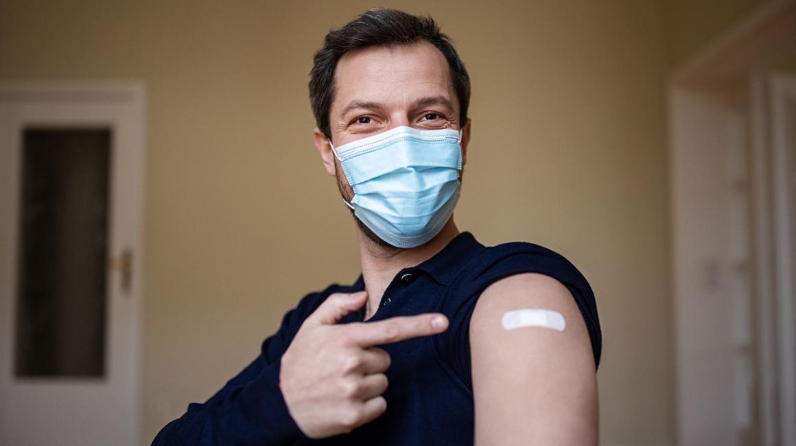 młody mężczyzna w maseczce pokazuje na ramię z plastrem po szczepieniu