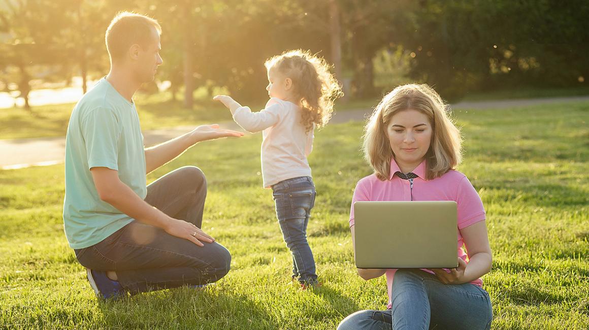 Ojciec bawi się z córeczką na łące, matka zagląda do laptopa
