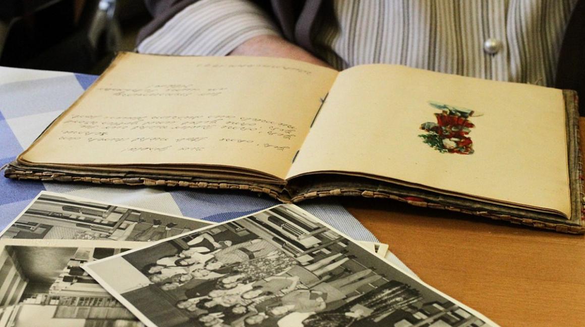 Leżące na stole lub biurku stare zdjęcia i otwarty pamiętnik. W tle zarys siedzącej osoby