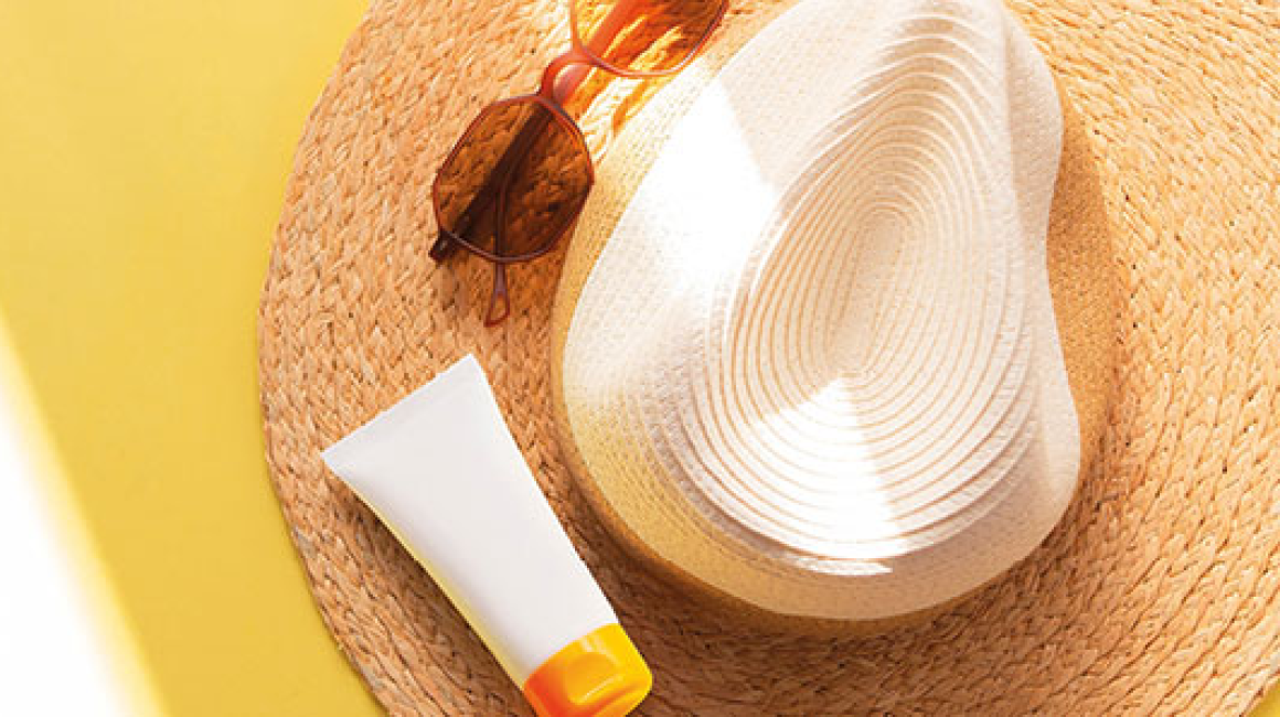 na żółtym słonecznym tle słomkowy kapelusz, na nim okulary i krem do opalania