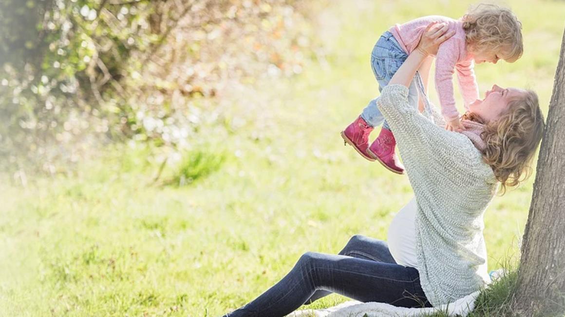 Kobieta w ciąży siedząca przy drzewie i trzymająca w górze dziecko