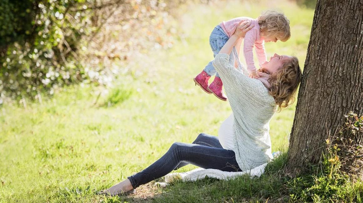 Kobieta w ciąży siedząca przy drzewie i trzymająca w górze dziecko