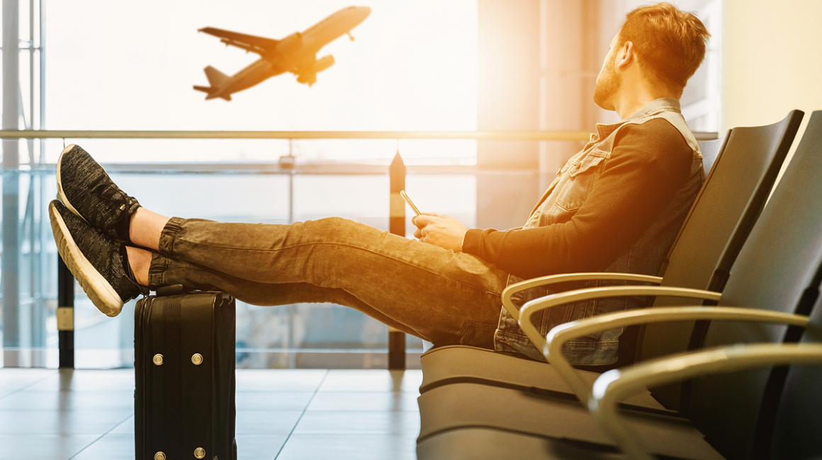 Mężczyzna siedzący w poczekalni lotniska. Uniesione nogi opiera na walice. Patrzy na widoczny za oknem startujący samolot
