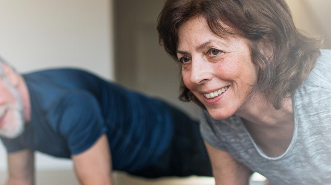 Starsza para ćwiczy w domu, twarzą do podłogi opierają się łokciami na macie