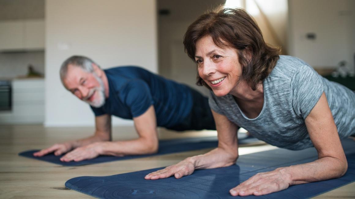 Starsza para ćwiczy w domu, twarzą do podłogi opierają się łokciami na macie