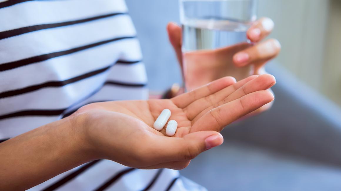 Wykadrowane ręce siedzącej osoby. W prawej ręce 2 tabletki, a w lewej szklanka z wodą