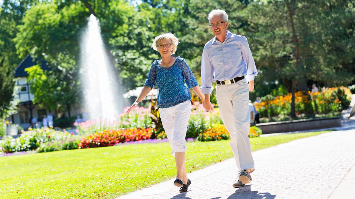 Para seniorów spacaruje po parku, w tle fontanna 