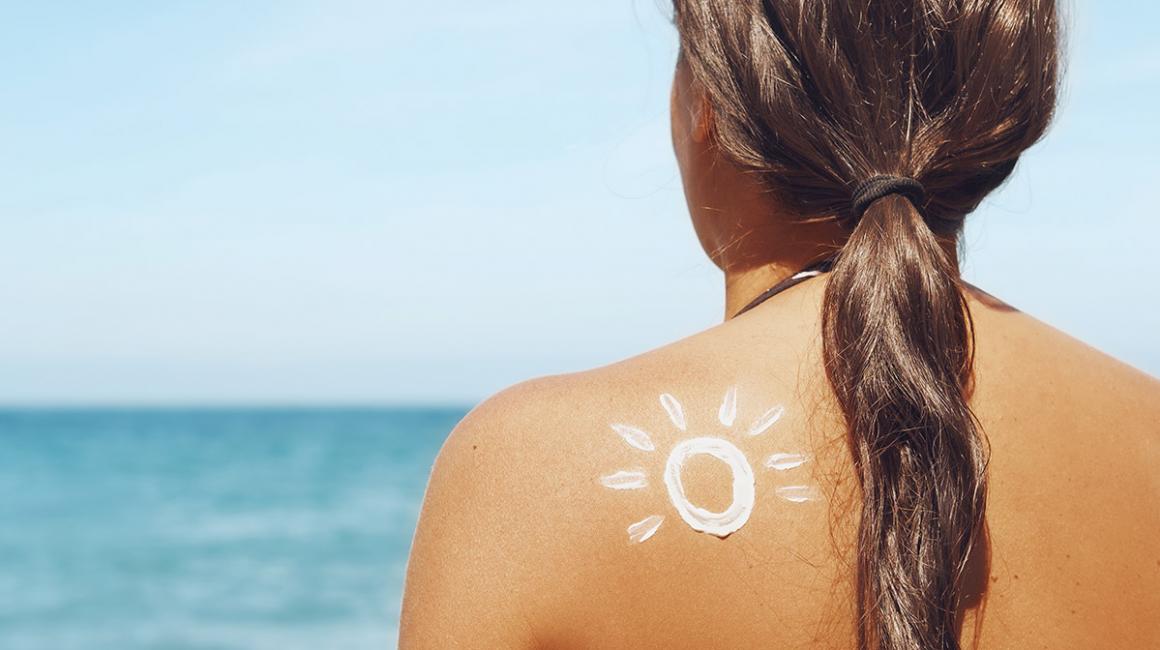 Kobieta stoi na plaży i patrzy na morze, widzimy jej opalone plecy posmarowane kremem z filtrem