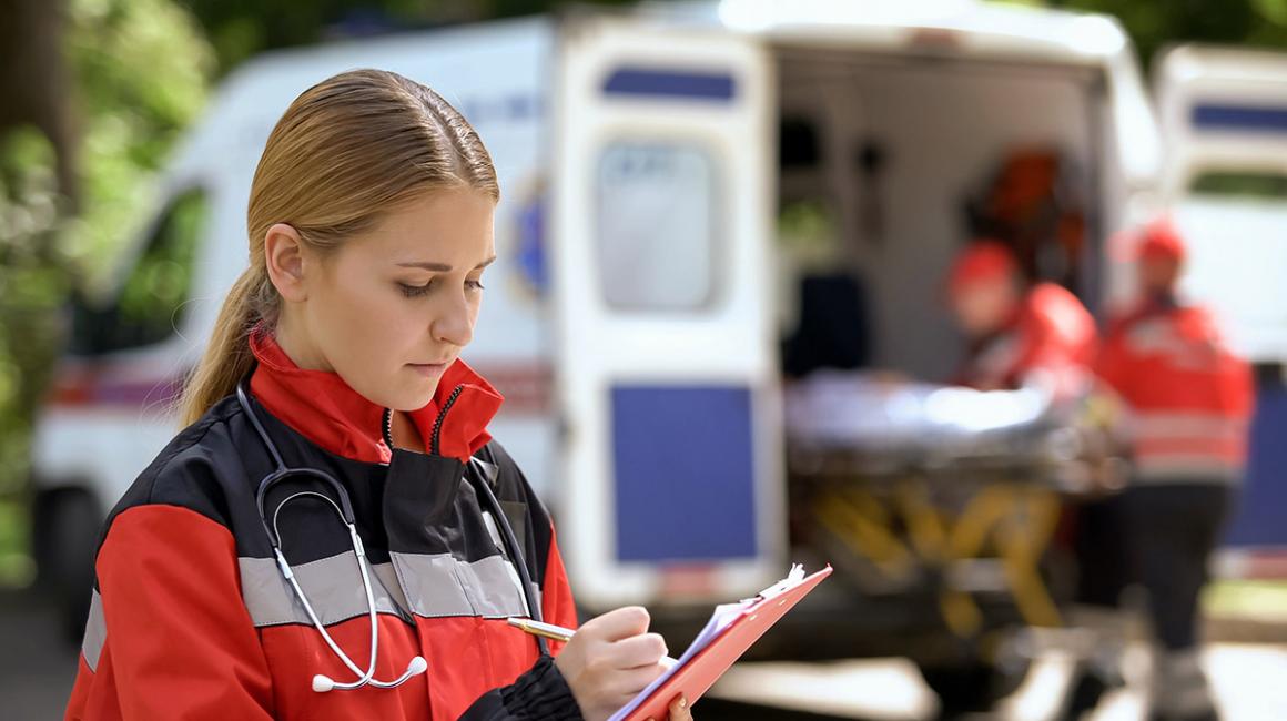 Na pierwszy planie kobieta zapisująca informacje w notesie. Na drugim planie karetka, z której 2 osoby wyjmują nosze.