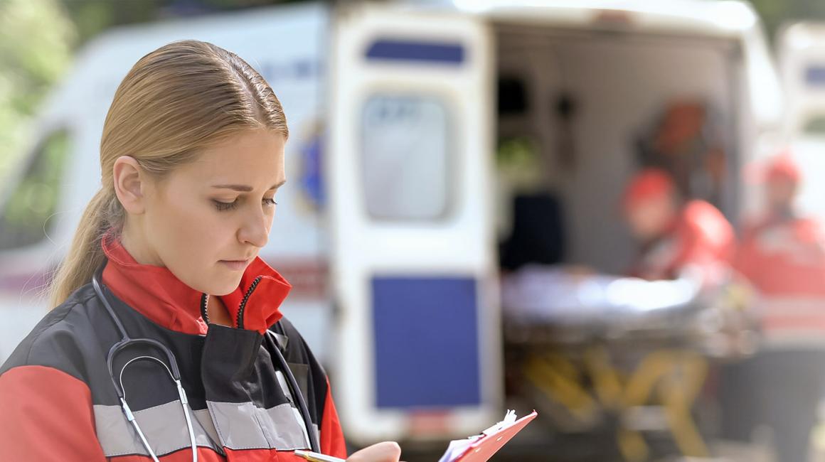 Na pierwszy planie kobieta zapisująca informacje w notesie. Na drugim planie karetka, z której 2 osoby wyjmują nosze.