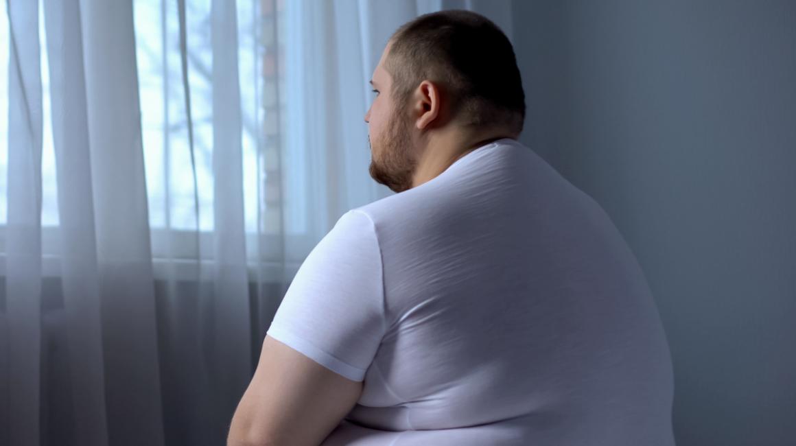 Otyły smutny mężczyzna siedzi na łóżku i patrzy w okno. 