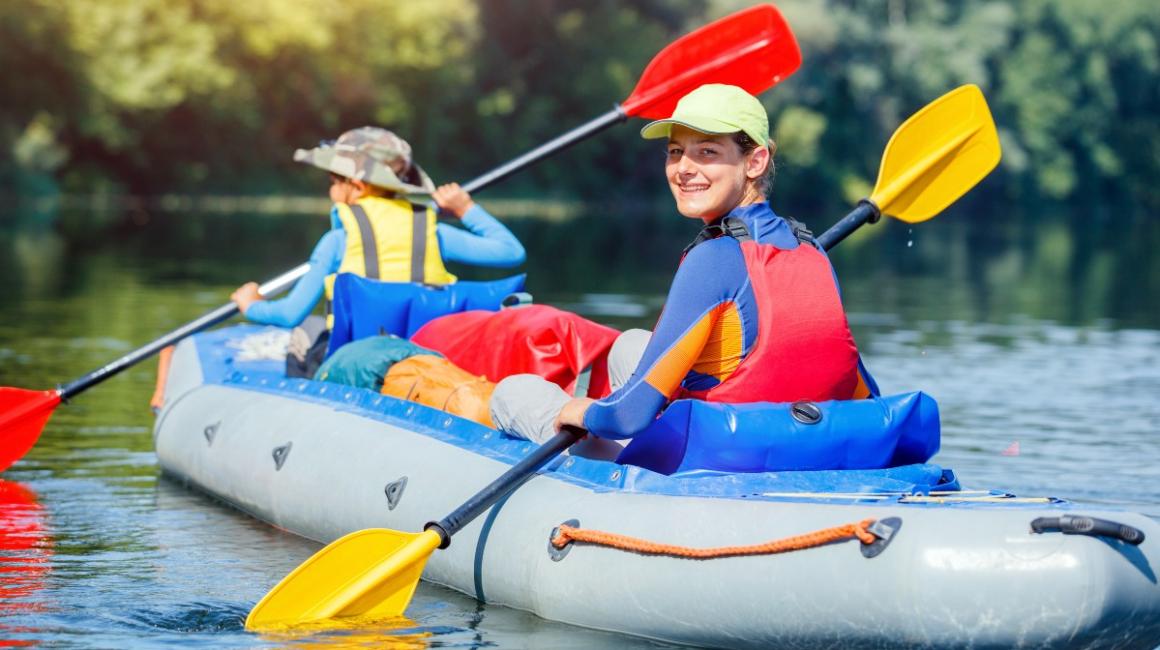 Uśmiechnięte dzieci ubrane w kamizelki ratunkowe płyną na rzece pontonem.