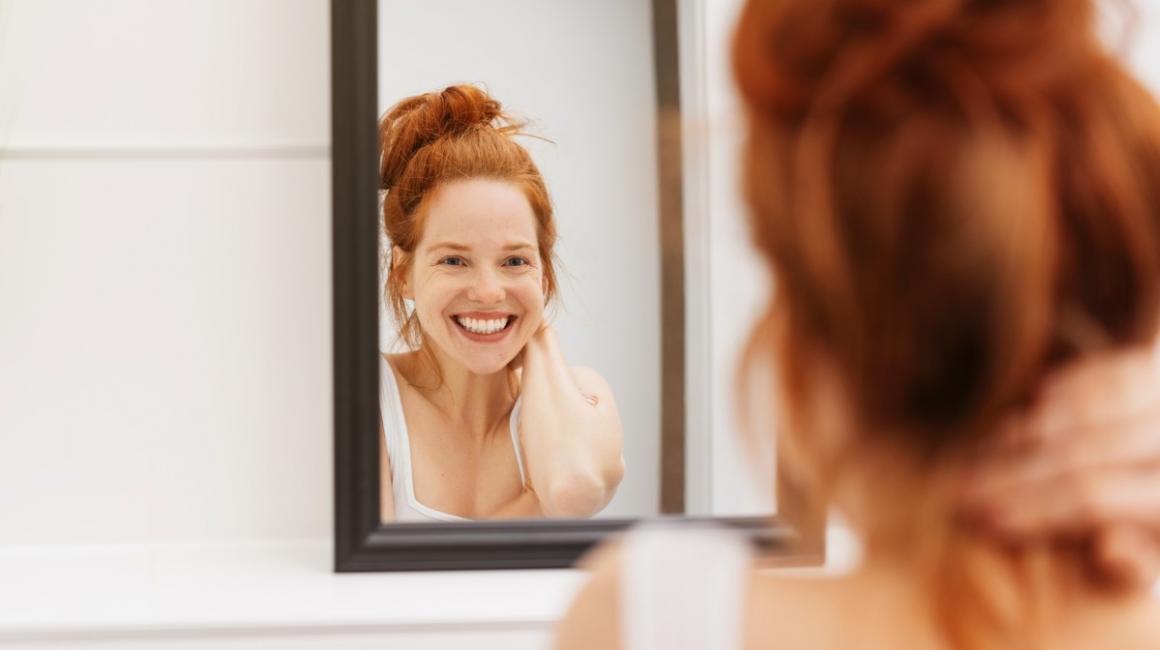 Uśmiechnięta kobieta stoi w łazience i przegląda się w lustrze.