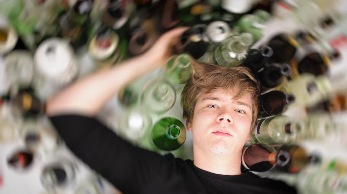 Nastolatek leży na podłodze, a dookoła niego stoją puste butelki po alkoholu.