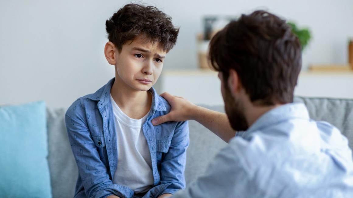 Trudna rozmowa ojca z synem. Chłopiec jest zasmucony, ojciec go wspiera, trzymając rękę na ramieniu.