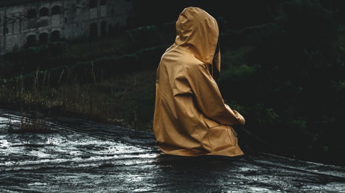 Siedząca tyłem na zaniedbanym wyłomie budynku osoba w żółtym płaszczu przeciwdeszczowym, z kapturem na głowie