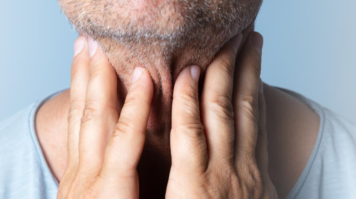 Mężczyzna dotyka palcami szyi. Na twarzy na grymas bólu. 