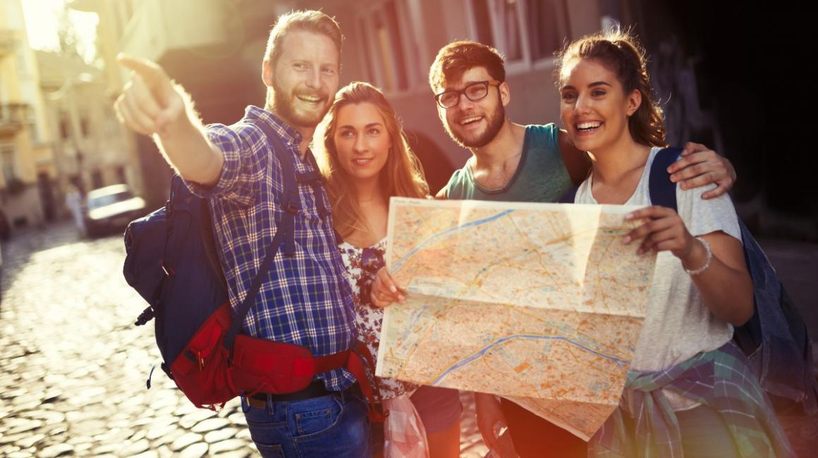 Czwórka uśmiechniętych młodych ludzi jest na wycieczce. Stoją na starej uliczce i trzymają dużą mapę. Mężczycna wskazuje kierunek palcem.