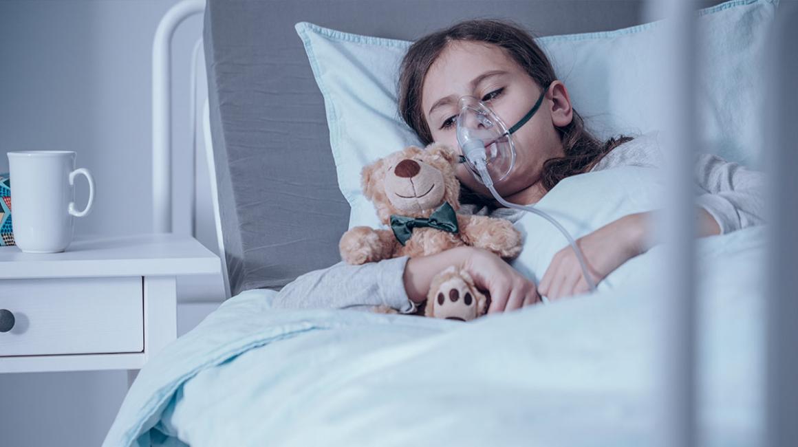 Dziecko w masce tlenowej na szpitalnym łóżku. Przytula misia.