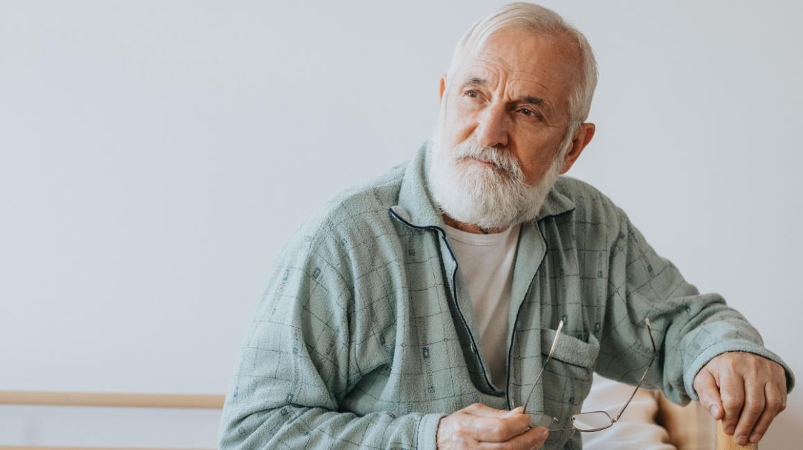 Starszy mężczyzna z siwymi włosami i brodą siedzi na łóżku i patrzy w dal.