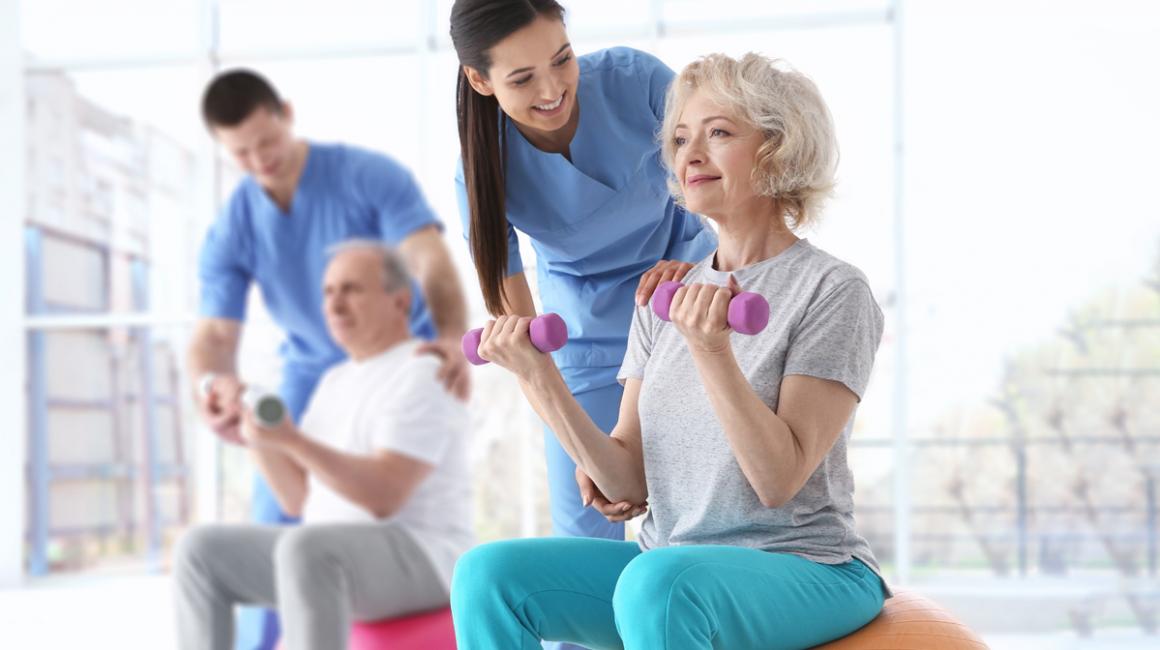 2 osoby ćwiczące na piłce i z cieżarkami na sali gimnastycznej pod ohiem rehabilitanta