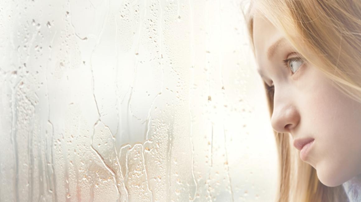 Smutna dziewcynka z długimi blond włosami patrzy przez szybę w dal. Za oknem pada deszcz.