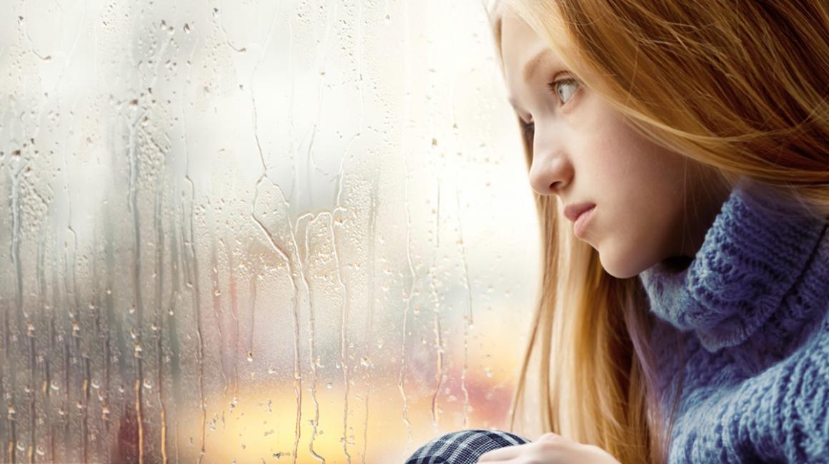 Smutna dziewcynka z długimi blond włosami patrzy przez szybę w dal. Za oknem pada deszcz.