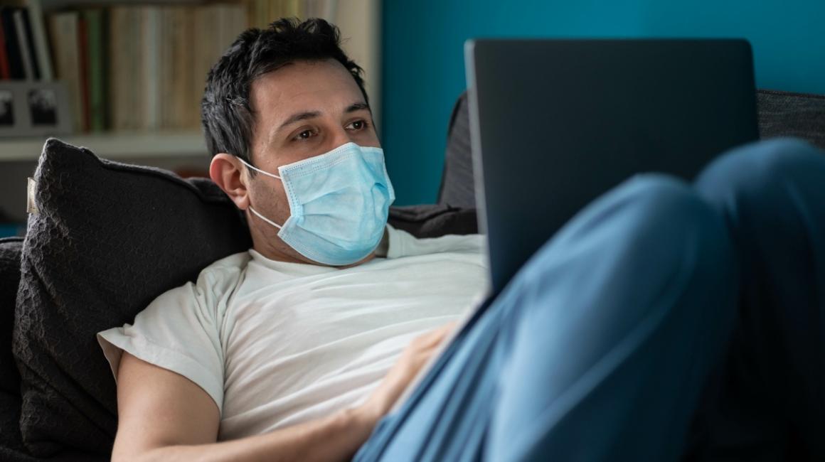 Mężczyna leży na kanapie z laptopem na kolanach i umawia się na test covid przez internet.