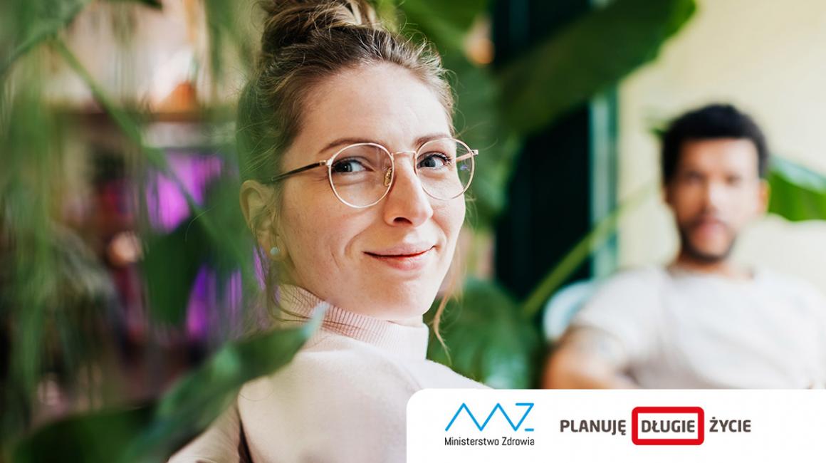 Odwrócona w stroną oglądającego siedzaca kobieta w okularach. Na drugim placie siedzący mężczyzna