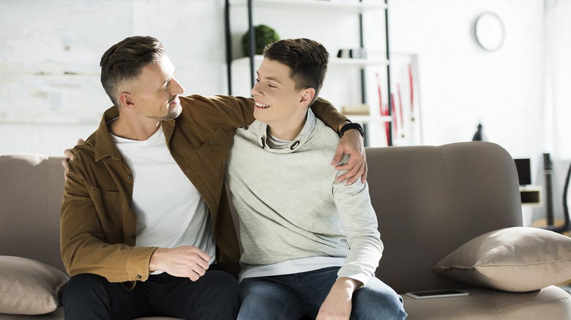 Ojciec obejmuje ramieniem nastoletniego syna, siedzą na kanapie