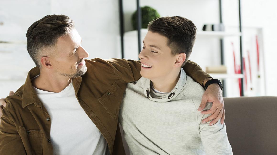 Ojciec obejmuje ramieniem nastoletniego syna, siedzą na kanapie
