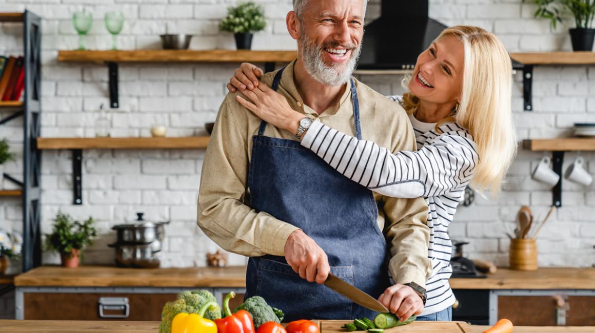 Kobieta i mężczyzna w kuchni. Mężczyzna kroi warzywa, kobieta obejmuje go za szyję.