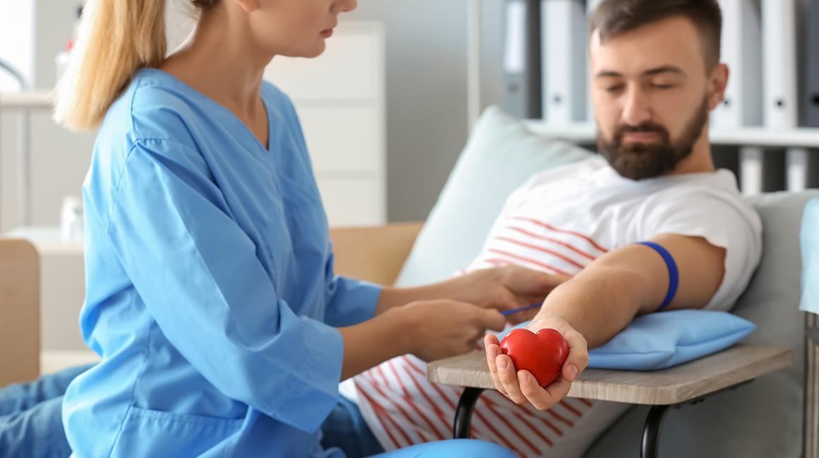 Pielęgniarka przygotowuje mężczyznę do oddania krwi. Zakłada mu opaskę uciskową. Mężczyzna trzyma w dłoni gumowe czerwone serce.