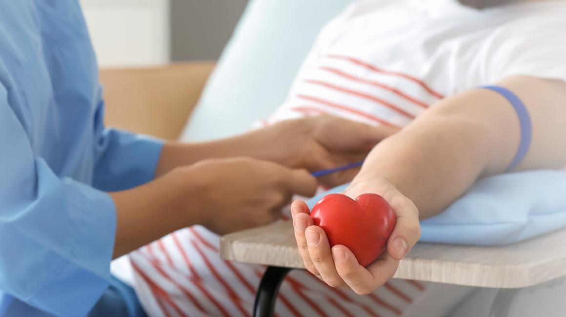 Pielęgniarka przygotowuje mężczyznę do oddania krwi. Zakłada mu opaskę uciskową. Mężczyzna trzyma w dłoni gumowe czerwone serce.