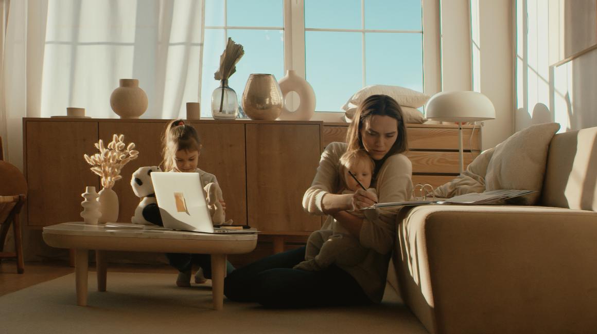 Kobieta siedzi na podłodze przy kanapie z małym dzieckiem, starsze dziecko bawi się na stoliku