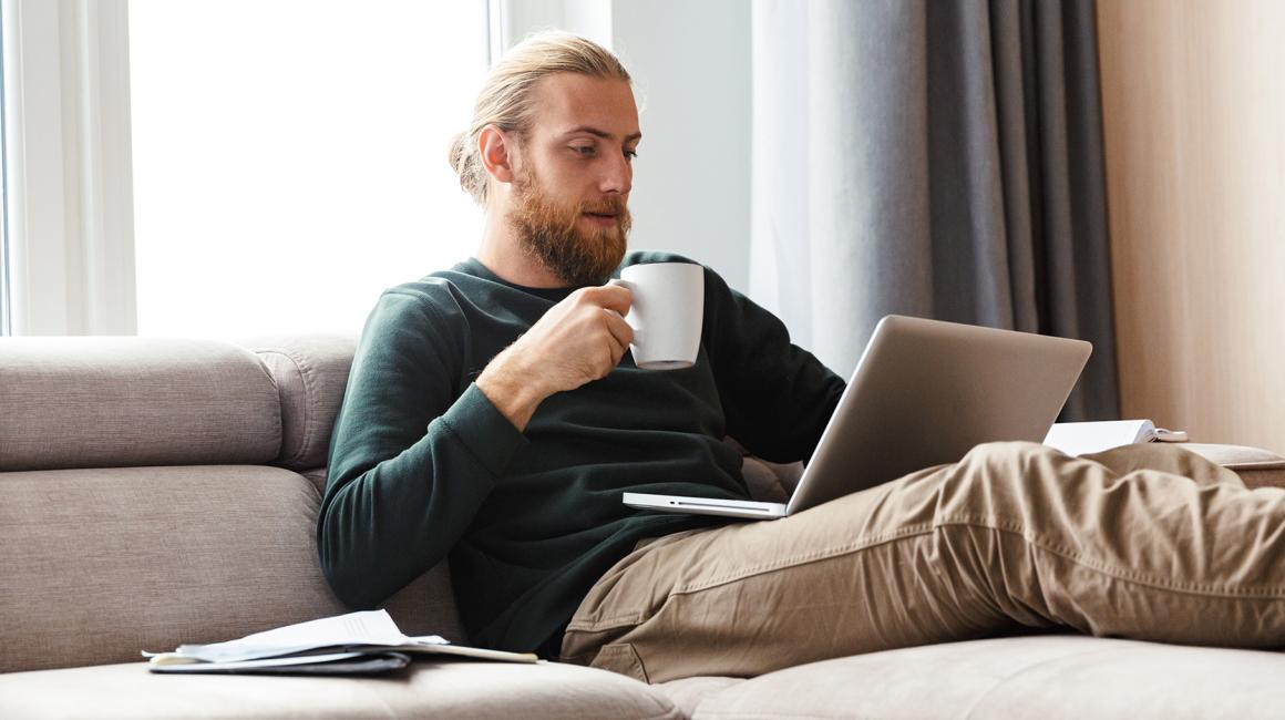 Brodaty młody facet z kubkiem na kanapie i laptopem na kolanach