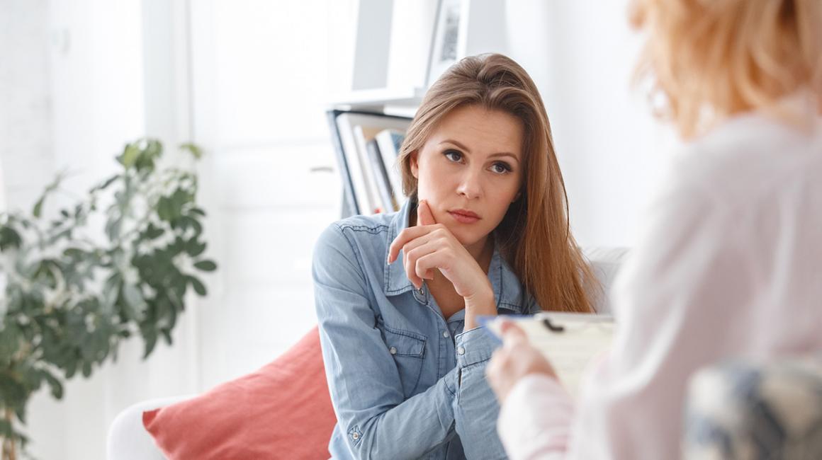 Na pierwszym planie mamy zadbaną młodą kobietę. Słucha, co mówi druga, która siedzi do nas tyłem.