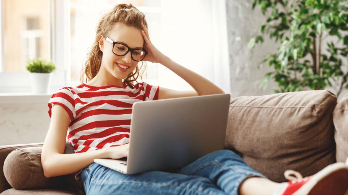 Młoda dziewczyna w okularach i koszulce w czerwone paski siedzi z laptopem na kolanach.