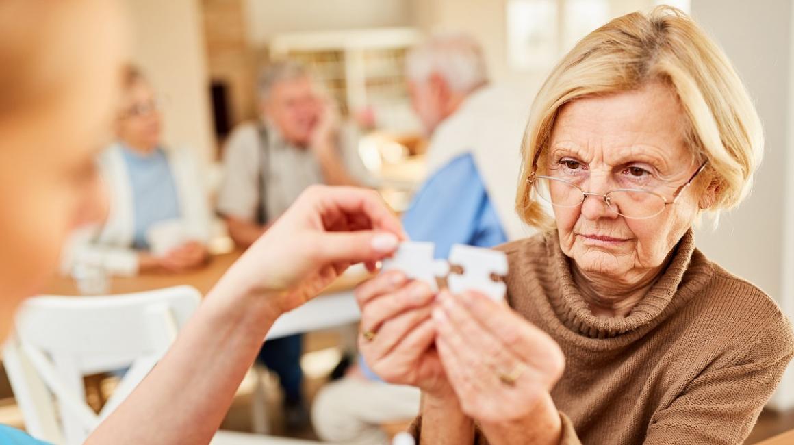 Starsza kobieta w okularach trzyma dwa puzzle i próbuje je dopaswać. Ktoś jej pomaga.
