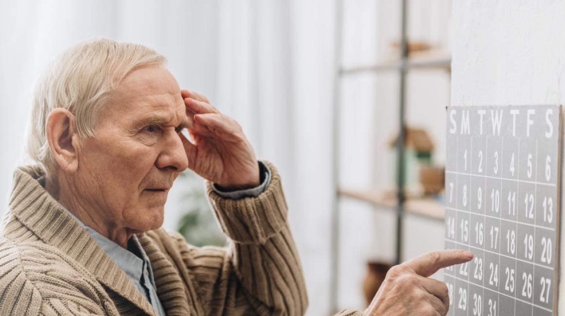 Starszy mężczyzna patrzy na kalendarz i wskazuje datę. Drugą ręką dotyka skroni. Wygląda na zdezorienrowanego.