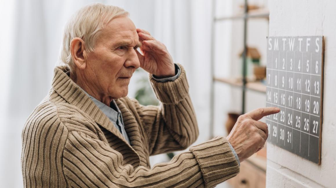 Starszy mężczyzna patrzy na kalendarz i wskazuje datę. Drugą ręką dotyka skroni. Wygląda na zdezorienrowanego.