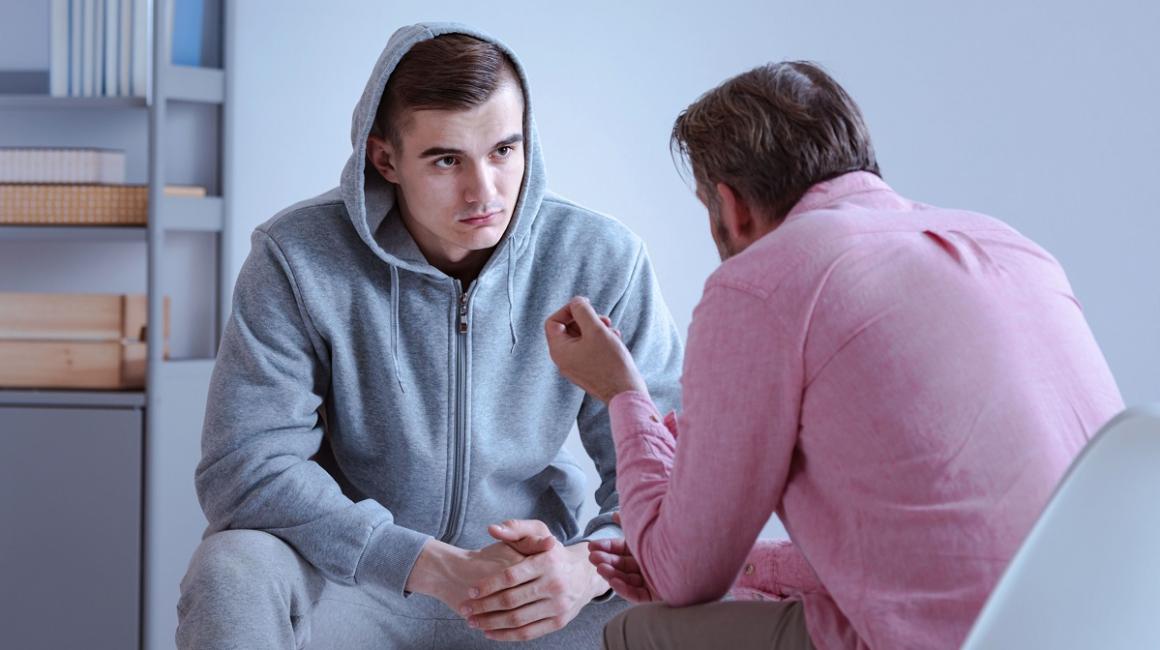 Młody chłopak siedzi i rozmawia z terapeutą. Ma na sobie dres, a na głowie kaptur.